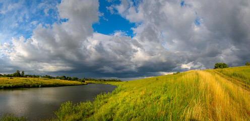 Summer rural panoramic view with calm river and dark stormy clouds over the farm fields
