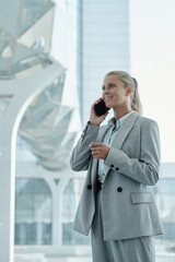 Happy young blond businesswoman in grey suit talking on mobile phone while standing by large iwndow in modern office