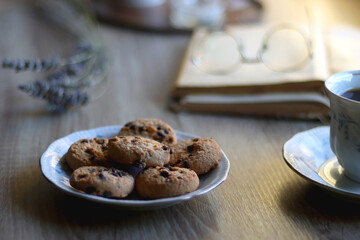Cup of tea, plate with chocolate chip cookies, open book, reading glasses, lit candles and dry lavender flowers. Hygge at home, selective focus.