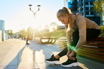 Young blond female athlete bending over leg bent in knee while sitting on wooden bench in urban environment in the morning