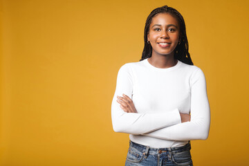 Well-looking lady with brunette curly hair looking at camera and smiles, holding arms crossed. Waist up portrait view of the charming smiling woman isolated on yellow background