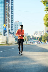 Front view of young African American athlete in red sport jacket, black leggiins, sneakers and baseball cap running along asphalt road