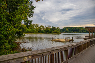 a gorgeous autumn landscape on Catawba River with a brown wooden dock over the rippling green waters surrounded by lush green trees and autumn colored trees with powerful clouds at sunset - Powered by Adobe