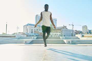 Fototapeta na wymiar Young active African American man in sportswear jumping over skipping rope while exercising on wide sunlit square in the morning