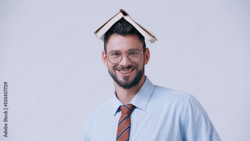 Wall mural cheerful teacher with book on head smiling at camera isolated on grey.