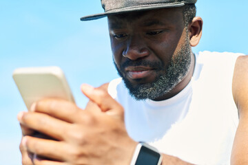 Close-up of face of young bearded African American sportsman in white t-shirt and black baseball cap scrolling in mobile phone outdoors