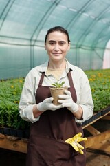 Ai generated face of gloved female fermer in apron holding pot with green seedling against workplace