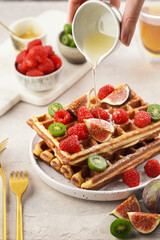 Pouring honey over fresh homemade sweet belgian waffles with powdered sugar with fresh raspberries, kiwi berries and figs on white plate