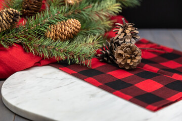 Red checkered kitchen towel, pine cones, ribbon, pine needles, pine branch, marble cutting board, Christmas concept, perspective view.