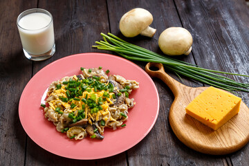 Pasta in a creamy sauce with mushrooms in a plate on a dark wooden table next to a glass of cream, cheese on the board, green onions and mushrooms.