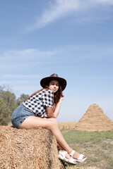 Naklejka na ściany i meble beautiful woman in plaid shirt and cowboy hat resting on haystack against sky background