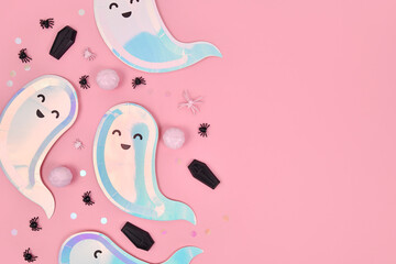 Cute pastel colored Halloween party flat lay with ghost shaped plates, coffins and confetti on pink background
