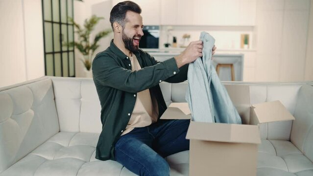 Handsome positive caucasian man opening big carton box while sits on sofa, unpacking internet store order at home, feel satisfied with fast delivery service and the goods which received, smile happily