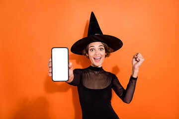 Portrait of delighted excited girl raise fist showing empty space telephone screen isolated on orange color background