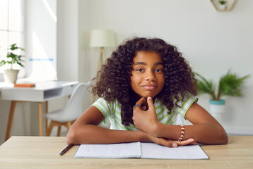 Portrait of happy school child at desk with notebook. Beautiful African American student girl sitting at table at home, looking at camera and smiling. Videocall, videoconference laptop PC screen view