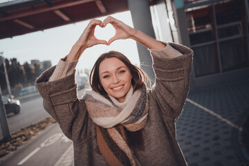 Photo of shiny charming lady dressed warm coat scarf rising arms showing heart enjoying sunny weather outdoors urban town road