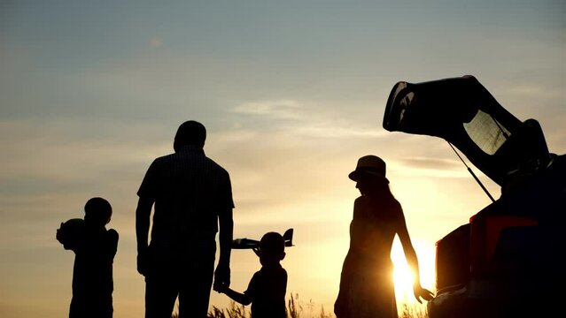 Family silhouettes spend time together in countryside near open car boot. Siblings holding parents hands play with toys at back sunset on blue sky