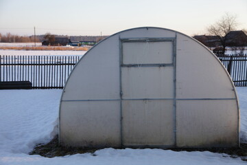 Polycarbonate arch greenhouse onn snowy country yard at winter day .