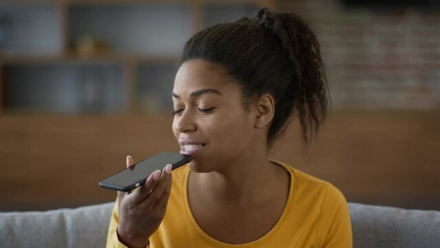 Modern communication concept. Young black woman recording audio message to friend, talking to phone mic at home