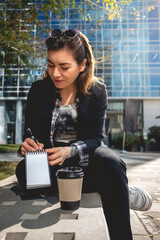 Young, successful and concentrated latina businesswoman with sunglasses and a cup of coffee taking notes of their ideas in a notebook in a park between skyscrapers