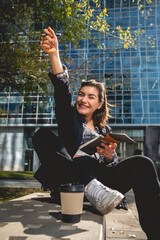 Fototapeta premium Young, successful and happy latina businesswoman with sunglasses and a cup of coffee taking notes of their ideas in a notebook in a park between skyscrapers