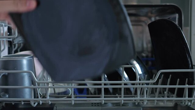 Person loads built-in dishwashing machine. Male hand places greasy black plates in first rack and basket of silver cutlery. Bright cups stand upside down