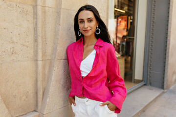 Stylish young woman relaxing outdoors during day. Brunette smiles looking away, wears pink shirt and white shorts. Stylish lady having good time.