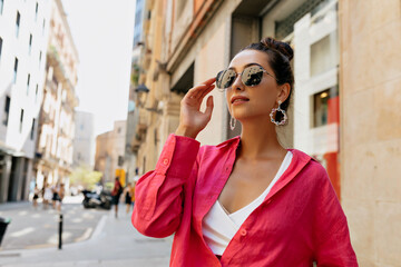 CLose up outdoor photo of elegant charming girl with dark hair wearing pink blouse and sunglasses looking aside and touching her glasses on summer street