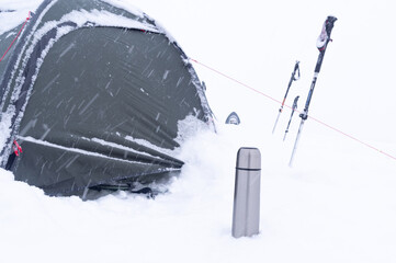Tourist tent in the snow. Thermos and trekking poles in the snow. Tourist equipment. Snowfall on a...