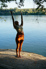 A yogi woman is doing sun salutation near the water on a dock at sunset.