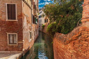 Antique red brick wall, brick walls of houses, green tree are reflected on the water surface of a narrow canal street in Venice, reflections of houses with windows and the sky on a Venetian street