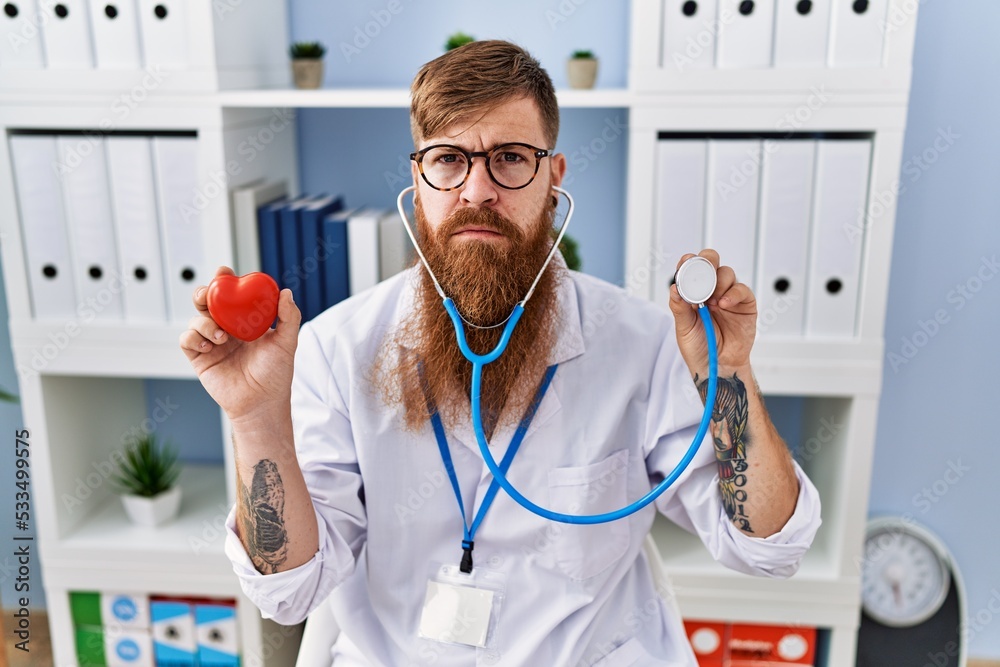 Canvas Prints redhead man with long beard wearing doctor uniform holding heart and stethoscope relaxed with seriou