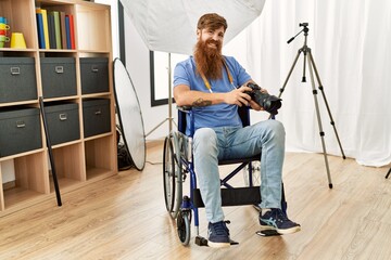 Fototapeta na wymiar Young redhead man photographer sitting on wheelchair using professional camera at photograpy studio