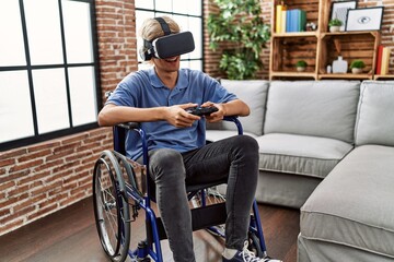 Fototapeta na wymiar Young blond man playing video game using virtual reality glasses and joystick sitting on wheelchair at home
