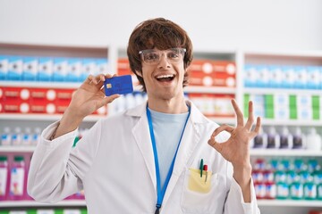 Hispanic young man working at pharmacy drugstore holding credit card doing ok sign with fingers, smiling friendly gesturing excellent symbol