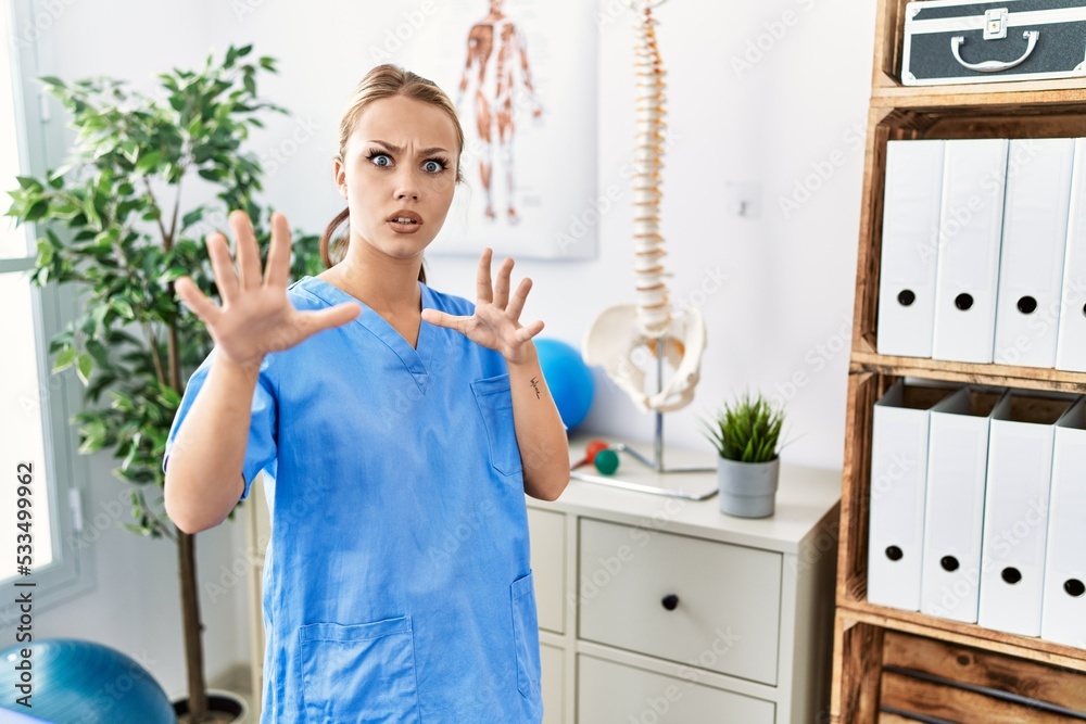 Sticker Young caucasian woman working at pain recovery clinic afraid and terrified with fear expression stop gesture with hands, shouting in shock. panic concept.