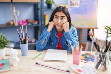 Young hispanic girl artist sitting on desk with doubt expression at art studio