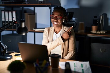 Beautiful black woman working at the office at night cheerful with a smile of face pointing with hand and finger up to the side with happy and natural expression on face