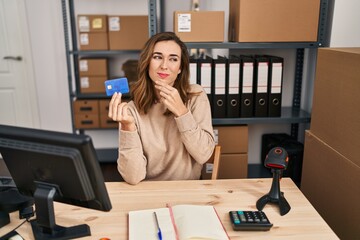 Young woman working at small business ecommerce holding credit card serious face thinking about...