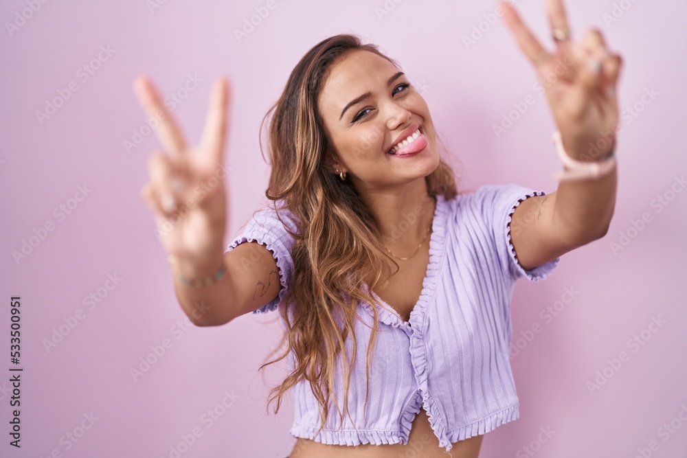 Wall mural Young hispanic woman standing over pink background smiling with tongue out showing fingers of both hands doing victory sign. number two.