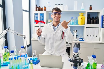 Young hispanic man working at scientist laboratory holding diploma pointing thumb up to the side smiling happy with open mouth