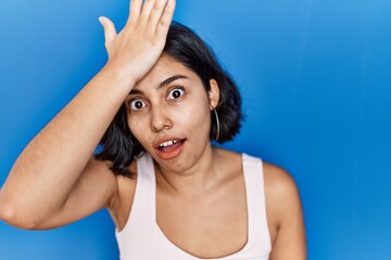 Young hispanic woman standing over blue background surprised with hand on head for mistake,...