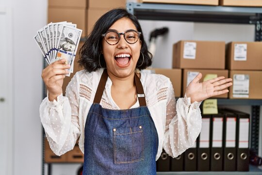 Young hispanic woman working at small business ecommerce holding dollars celebrating achievement with happy smile and winner expression with raised hand