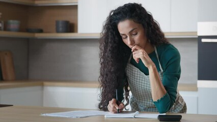 Concentrated hispanic woman wife young mom stand in modern kitchen make notes at copybook write recipe grocery list calculate domestic utilities use calculator count bills puzzled by monthly expenses