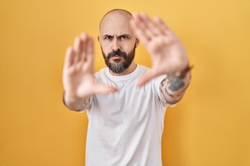 Young hispanic man with tattoos standing over yellow background doing frame using hands palms and fingers, camera perspective