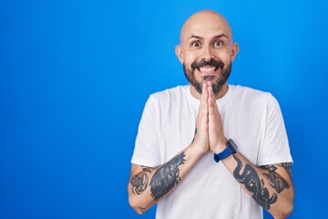 Hispanic man with tattoos standing over blue background praying with hands together asking for forgiveness smiling confident.