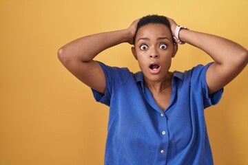 African american woman standing over yellow background crazy and scared with hands on head, afraid and surprised of shock with open mouth