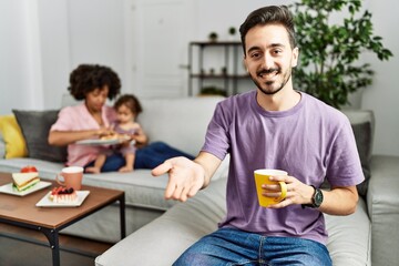 Hispanic father of interracial family drinking a cup coffee smiling cheerful offering palm hand giving assistance and acceptance.