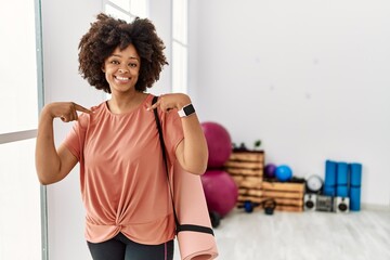 Fototapeta na wymiar African american woman with afro hair holding yoga mat at pilates room looking confident with smile on face, pointing oneself with fingers proud and happy.