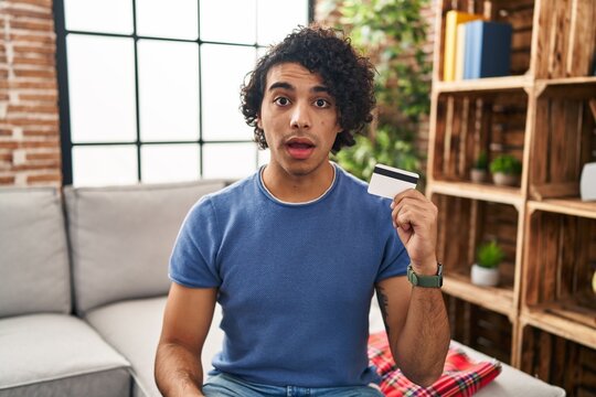 Hispanic man with curly hair holding credit card scared and amazed with open mouth for surprise, disbelief face